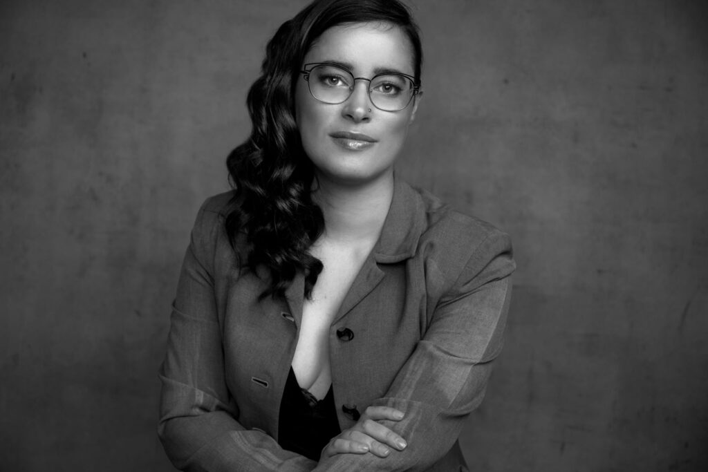 A black and white portrait of Saro Lynch-Thomason. Her face and upper body are visible. She rests her right hand on her left elbow and looks directly out at the viewer. She appears calm and confident. She is wearing an unbuttoned jacket with a black shirt underneath. 